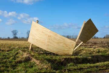 Angled plywood panels create an unusual structure in a sunny field, surrounded by dry grass and distant trees under a bright blue sky.