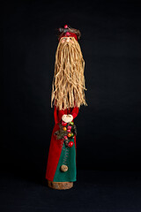 Handcrafted Christmas figure with a long straw beard, red and green velvet robes, and festive berry accents. A traditional holiday decoration standing on a dark background.