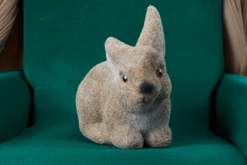 Adorable fuzzy brown bunny figurine with bright red eyes sits comfortably on a soft dark green upholstered chair, evoking warmth and charm.