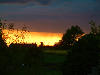 Obraz premium Sunset over a rural landscape with silhouetted trees and buildings.