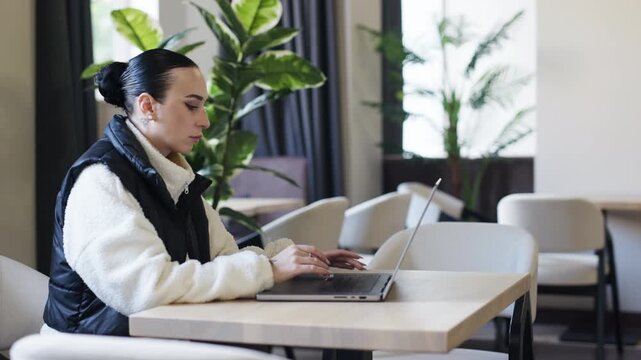 Young woman with dark hair, wearing a cozy sweater and vest, is focused on her laptop in a modern coworking space, surrounded by greenery, embodying remote work lifestyle	