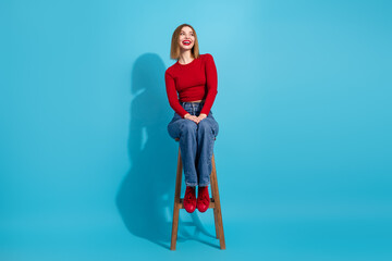 Lovely girl in fashionable red top posing on wooden stool against a bright blue background, cheerful and adorable