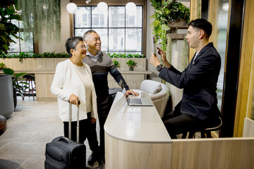 Happy senior Asian couple checking in at hotel on holiday vacation with young male receptionist....
