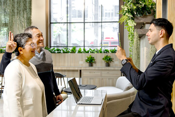 Happy senior Asian couple checking in at hotel on holiday vacation with young male receptionist....