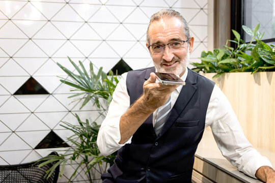 Happy senior businessman talks on smartphone at modern office. Elderly man connects with AI technology at co working space office workplace. Mature manager uses mobile phone to control remotely.