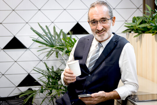 Happy senior businessman drinking coffee and holding smartphone at modern office. Elderly man sipping coffee at co working space office workplace during a break. Mature manager taking a break.