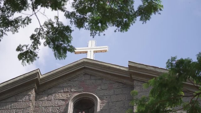 Timelapse of San Juan de Dios Church in San Rafael, Bulacan, Philippines, featuring the historic stone facade and cross under a bright sky framed by green tree branches, showcasing heritage architectu