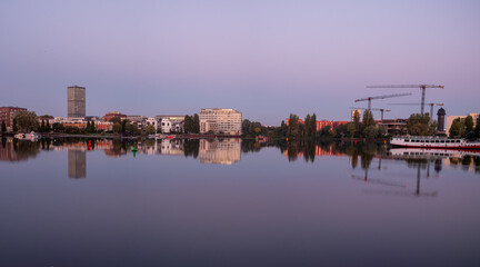 Naklejka premium Berlin Cityscape at rummelsburg lake reflected in calm water at dusk, with modern buildings, cranes, and a boat under a purple evening sky