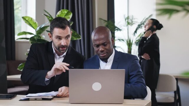Two professional men engaged in a collaborative discussion while working on a laptop at a modern office space, showcasing teamwork and innovation in a business environment	