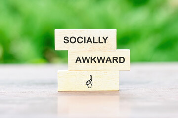 Wooden blocks are stacked. They spell out socially awkward and show a pointing finger. The background is a blurred green suggesting a garden