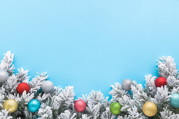 Colorful baubles with glitter and green artificial fir branches covered with hoarfrost and white snow. Pastel blue table background. Closeup. Christmas decoration. Empty place for text. Top down view.