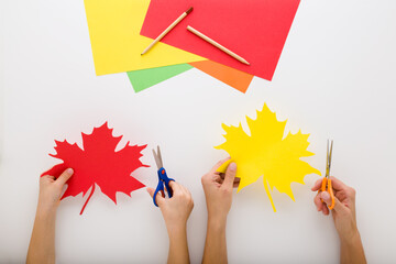 Young adult mother and little child hands cutting colorful maple leaves from application paper on light gray table background. Closeup. Making beautiful autumn decoration. Flat lay. Top down view.
