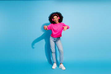 Trendy young woman in pink sweater and jeans enjoying music with headphones, expressing joy against bright blue background