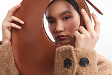 Obraz premium asian woman peers through a circular brown leather accessory, wearing a cozy textured coat. The close-up captures her curious, composed expression, delivering a stylish, artful mood.