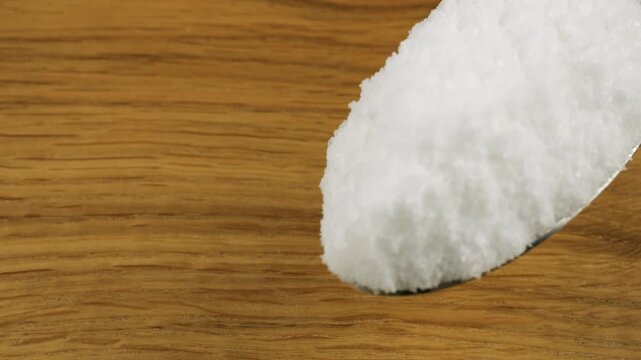 A tablespoon of fine crystals of natural sea salt is sprinkled onto a wooden surface. Close-up view.