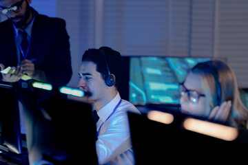 Dynamic night shift collaboration. Male manager provides guidance to a focused male colleague wearing a headset at his computer in a modern tech office during an intense work session.