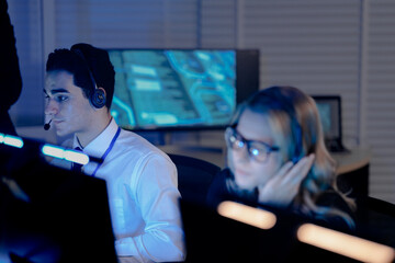 Dynamic night shift collaboration. Male manager provides guidance to a focused male colleague wearing a headset at his computer in a modern tech office during an intense work session.