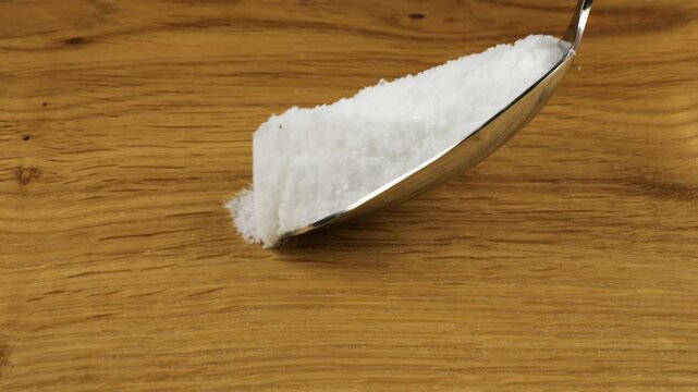 A tablespoon of fine crystals of natural sea salt is sprinkled onto a wooden surface. Close-up view.