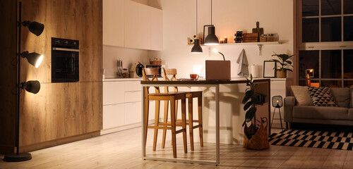 Interior of modern kitchen with counters, table and glowing lamps at night