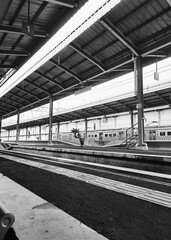 A train station in black and white. A train is seen waiting on a distant platform at this...