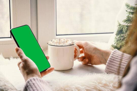 Close-up photo of a person holding a smartphone with a green screen chroma key near a window, holding a hot mug of cocoa or coffee, cozy winter setting, cold weather mockup, application display, casua