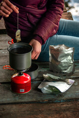 Traveler making hot drink outdoors with camping stove and freeze-dried food, adventure lifestyle near lake