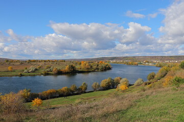 Peaceful autumn landscape with a calm river, colorful trees, and a grazing cow on a green hillside under a cloudy sky