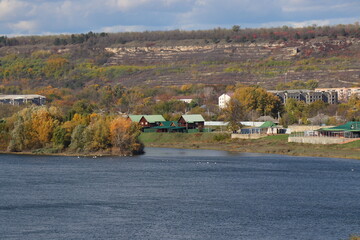 Peaceful autumn landscape with a calm river, colorful trees, and a grazing cow on a green hillside under a cloudy sky