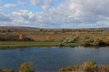 Peaceful autumn landscape with a calm river, colorful trees, and a grazing cow on a green hillside under a cloudy sky