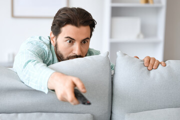 Scared young man with remote controller watching TV behind sofa at home