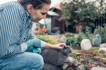 A young Caucasian woman with brown hair kneels in a garden, gently petting a gray cat. The scene...