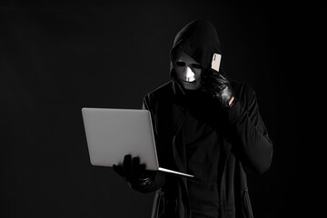 Male fraudster in mask with laptop talking by mobile phone on black background