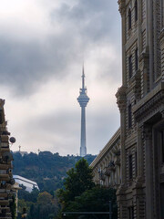 Dramatic cityscape featuring a towering communications tower piercing through a cloudy sky, flanked by grand classical buildings and lush green hills