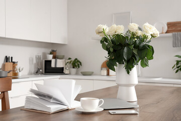 Vase with white roses, gadgets, coffee cup and book on table in kitchen