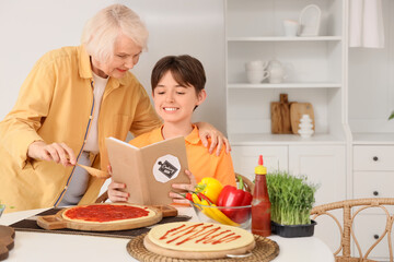 Teenage boy with recipe book and his grandmother making pizza in kitchen