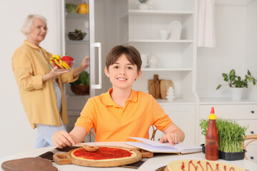 Teenage boy with recipe book and his grandmother making pizza in kitchen