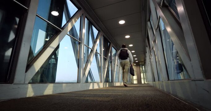 Low-angle wide slow-motion shot of male traveler with backpack and laptop bag hurrying and running along empty airport jet bridge toward airplane. Final call and last-minute boarding scene