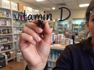 a person"s hand writes "vitamin D" with a marker in front of shelves stocked with health and wellness products inside a pharmacy.
