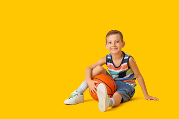 Stylish little boy with ball sitting on yellow background