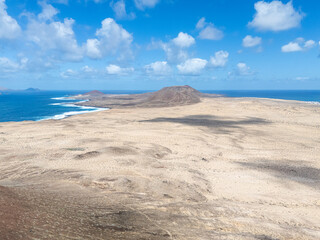 Aerial view of a volcanic cone rising from the arid landscape of La Graciosa Island, Canary Islands, with the Atlantic Ocean in the background.
