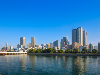 Fototapeta premium Modern city skyline of skyscrapers and a bridge viewed across a river under a clear blue sky (viewed from Hamarikyu Garden, Tokyo, Japan)