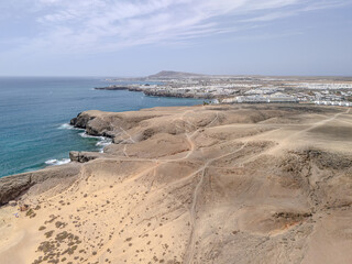 Aerial view of rugged cliffs and golden sand beach near Playa Blanca, Lanzarote, with turquoise Atlantic waters and arid volcanic terrain.
