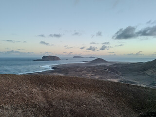 Aerial view of a volcanic cone rising from the arid landscape of La Graciosa Island, Canary Islands, with the Atlantic Ocean in the background.
