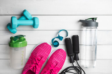 Top view of pink sneakers, blue dumbbells, jump rope water bottle, shaker and measuring tape on white wooden background with copy space, fitness equipment flat lay concept