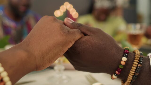 Extreme close-up shot of unknown man and woman wearing stone bracelets joining hands praying before festive Kwanzaa lunch
