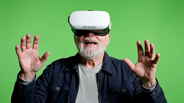 An amazed senior man with a white beard wearing a VR headset raises his hands in surprise experiencing immersive virtual reality for the first time against a green screen - Powered by Adobe