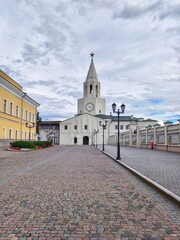Obraz premium The Spasskaya Tower, main gate of the Kazan Kremlin,a white tower with a clock on it and a tall pointed spire topped with a star and the walkway in front with old-style street lamps lining the sides 