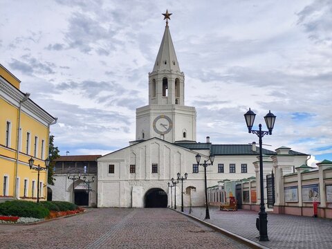 The Spasskaya Tower, main gate of the Kazan Kremlin,a white tower with a clock on it and a tall pointed spire topped with a star and the walkway in front with old-style street lamps lining the sides