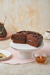 A homemade Sachertorte made of two chocolate layers, layered with apricot jam and topped with icing, on a white cake stand against a light concrete background.