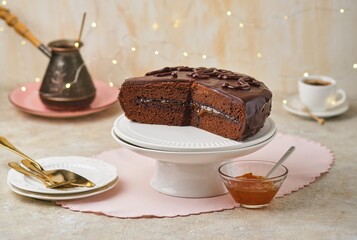A homemade Sachertorte made of two chocolate layers, layered with apricot jam and topped with icing, on a white cake stand against a light concrete background.